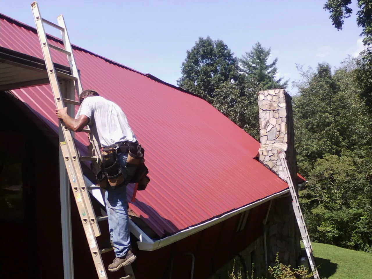Roofer on a ladder beside a red metal roof and stone chimney — Soffit & Fascia Repair by Roofing Brothers in Blue Mound